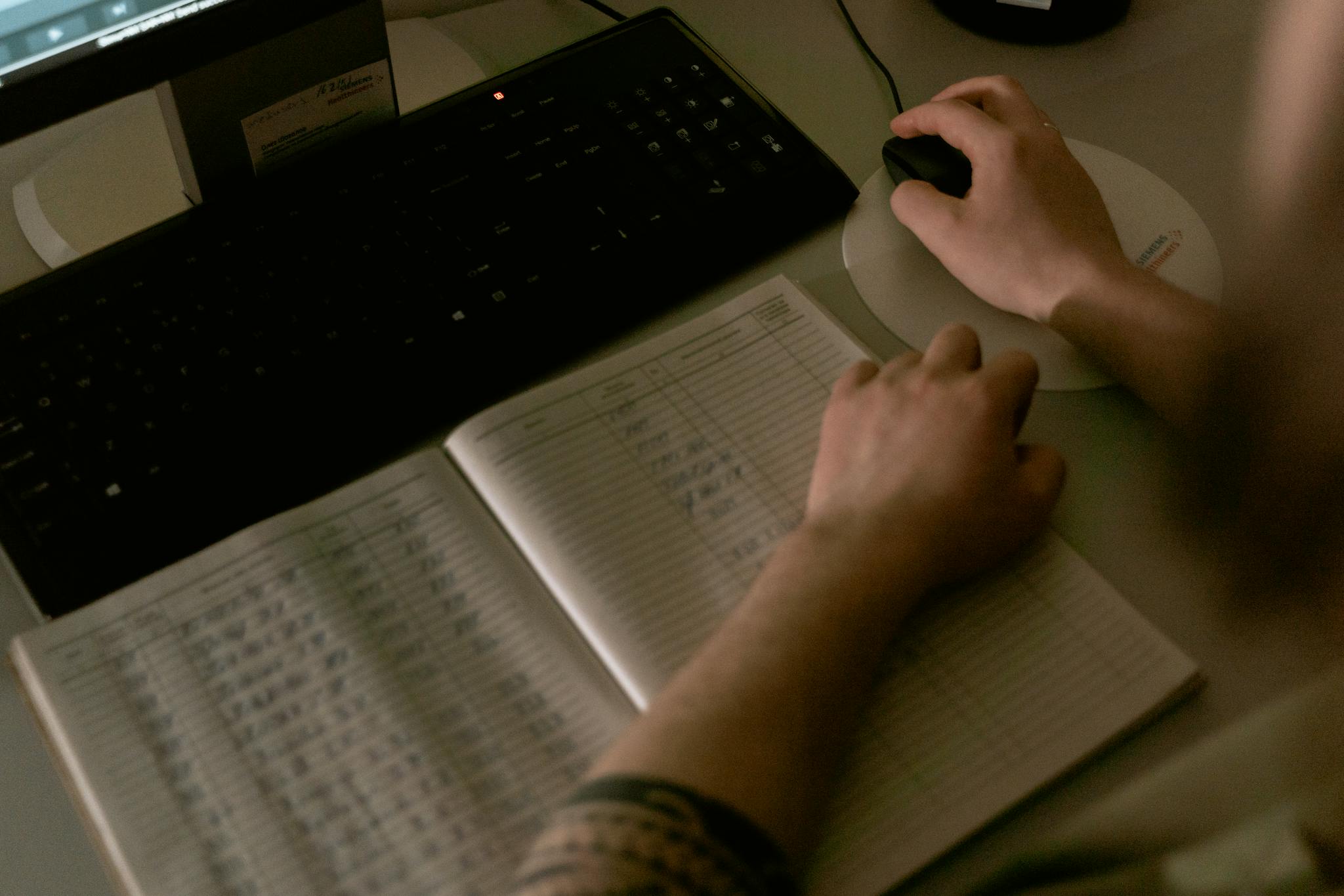 Healthcare worker reviewing medical data on a computer with a notebook open with written details, in a clinic setting.